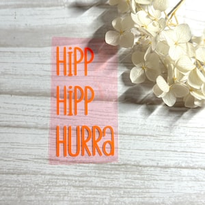 Könnte beinhalten: Orangefarbene Vinylbuchstaben auf rosa Untergrund. Der Schriftzug lautet "Hipp Hipp Hurra!"