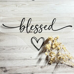 Könnte beinhalten: Ein klares Acrylschild mit dem Wort "blessed" in schwarzer Kursivschrift. Unter dem Wort befindet sich ein kleines schwarzes Herz. Das Schild befindet sich auf einem hellen Holzhintergrund.