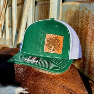 May include: Green and white trucker hat with a leather patch featuring a four-leaf clover design. The hat has a curved brim and a mesh back. The patch has the text "4-H" and "EST. 1902".