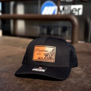 May include: Black trucker hat with a mesh back and a curved brim. The hat features a rectangular leather patch with the word "Pipeline" and the text "Since 1865" in a decorative font. The patch has an ornate floral design.