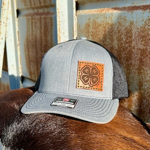 Puede incluir: Gorra trucker gris con parte trasera de malla negra y visera curva. La gorra presenta un parche cuadrado de cuero marrón con un diseño de trébol de cuatro hojas y las letras "4-H". Cierre a presión y etiqueta Richardson 112.