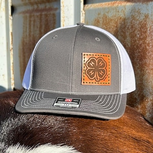 Puede incluir: Gorra de camionero gris y blanca con un parche de cuero con un diseño de trébol de cuatro hojas. La gorra tiene una visera curva y una parte trasera de malla. El parche tiene las letras "4-H" y las palabras "EST. 1970".