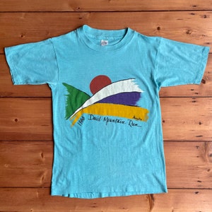 May include: A light blue t-shirt with a graphic design of a sunset over mountains. The text "1980 Devil Mountain Run" is printed on the shirt.