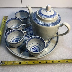 May include: A white ceramic tea set with blue patterns, including a teapot, four cups, and a round tray. A yellow measuring tape shows the tray is approximately 25.4 cm wide. The pieces feature translucent rice grain patterns and blue dragon motifs.