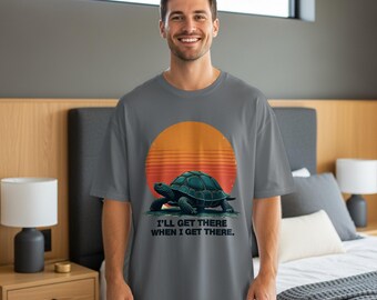 May include: A gray t-shirt featuring a turtle graphic with a sunset backdrop. The text "I'LL GET THERE WHEN I GET THERE" is printed below the turtle. The shirt is a relaxed fit.