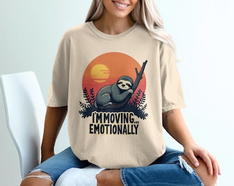 May include: Beige t-shirt featuring a sloth graphic. The sloth is perched on a branch against a sunset backdrop, with the text "I'M MOVING... EMOTIONALLY" below. The shirt is a relaxed fit.