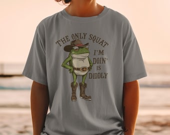 May include: A light gray t-shirt featuring a cartoon frog in a cowboy hat and boots. The frog has its hands on its hips and the text "THE ONLY SQUAT I'M DOIN' IS DIDDLY" is printed above the frog.