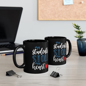 May include: Two black coffee mugs with white text that reads "my students stole my heart" with a red heart. The mugs are on a wooden table with a laptop, pencil, and paper clips.