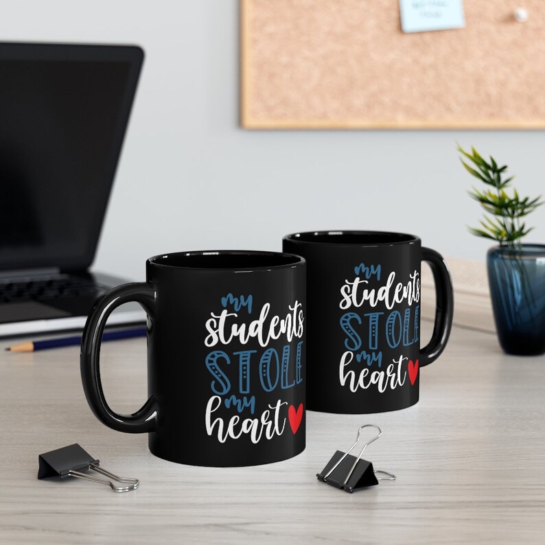 May include: Two black coffee mugs with white text that reads "my students stole my heart" with a red heart. The mugs are on a wooden table with a laptop, pencil, and paper clips.