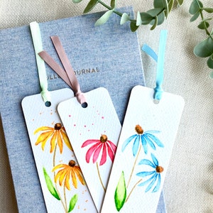 May include: Three watercolor bookmarks featuring different colored flowers. The bookmarks are white with a light brown ribbon attached to each. The flowers are painted in shades of yellow, pink, and blue.