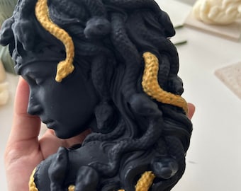 May include: Black and gold Medusa bust sculpture with snakes in her hair. The sculpture is made of resin and has a matte finish.