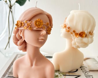 May include: Two decorative candles shaped like a woman's bust with a floral design covering the eyes. The candles are made of a light pink and white wax.