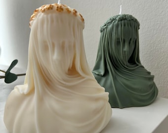 May include: Two decorative candles shaped like a bust of a person wearing a veil. The candle on the left is white with a gold crown, and the candle on the right is green.