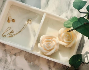 Op de afbeelding: Een wit marmeren dienblad met twee witte roosvormige kaarsen en een gouden ketting met een hartvormige hanger.