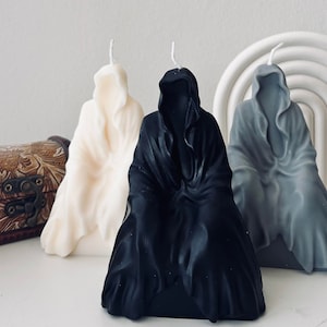 Puede incluir: Tres figuras de velas con capucha, una blanca, una negra y una gris, están dispuestas sobre una superficie blanca. Las velas tienen forma de figuras encapuchadas con las capuchonas levantadas.