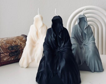 May include: Three hooded candle figures, one white, one black, and one gray, are arranged on a white surface. The candles are shaped like cloaked figures with their hoods pulled up.