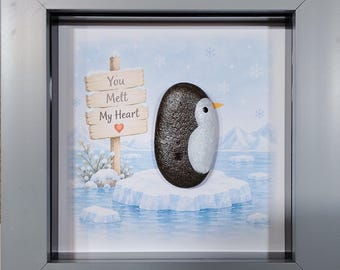 May include: A framed wall art piece featuring a smooth, dark brown and white pebble shaped like a penguin standing on an ice floe. The background illustration shows a snowy landscape with a wooden sign that reads You Melt My Heart with a red heart.