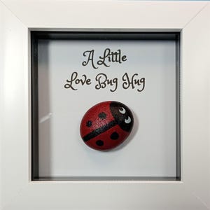 May include: A framed artwork featuring a painted ladybird on a white background. The ladybird is red with black spots and a black head. The text "A Little Love Bug Hug" is above the ladybird. The frame is white.