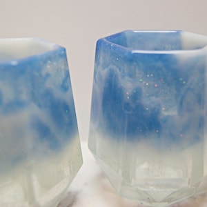 May include: Two clear glass cups with a blue and white marbled design. The cups have a geometric shape with a flat bottom and a wide opening.