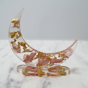 May include: A crescent moon shaped ring holder made of clear resin with pink pressed flowers and gold leaf accents. The holder sits on a small, clear resin base.