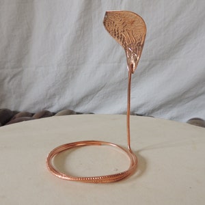 Könnte beinhalten: Eine Skulptur aus Kupferdraht, die eine Schlange mit einem strukturierten Kupferblatt auf der Oberseite darstellt. Die Schlange ist zu einem Kreis gewickelt und das Blatt steht aufrecht.