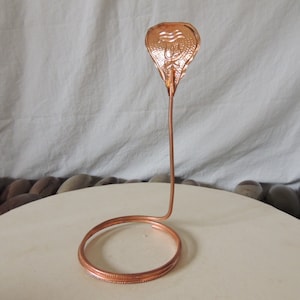 Könnte beinhalten: Eine Kupferschlangen-Skulptur mit einem gewundenen Sockel und einem flachen, strukturierten Kopf. Der Kopf hat ein erhabenes Design.