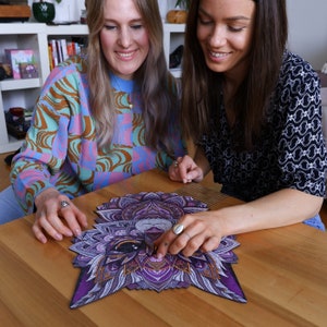 May include: Two people are working on a purple and gold mandala jigsaw puzzle of a cat's face. The puzzle is almost complete.