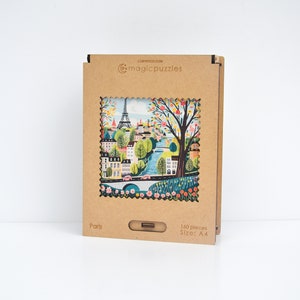 Może przedstawiać: Brązowe pudełko kartonowe z wyciętym obrazkiem kolorowej ilustracji wieży Eiffla i panoramy Paryża. Na pudełku nadrukowano tekst "magicpuzzles" i "Paris". Pudełko zawiera 180 element&oacute;w i ma rozmiar A4.