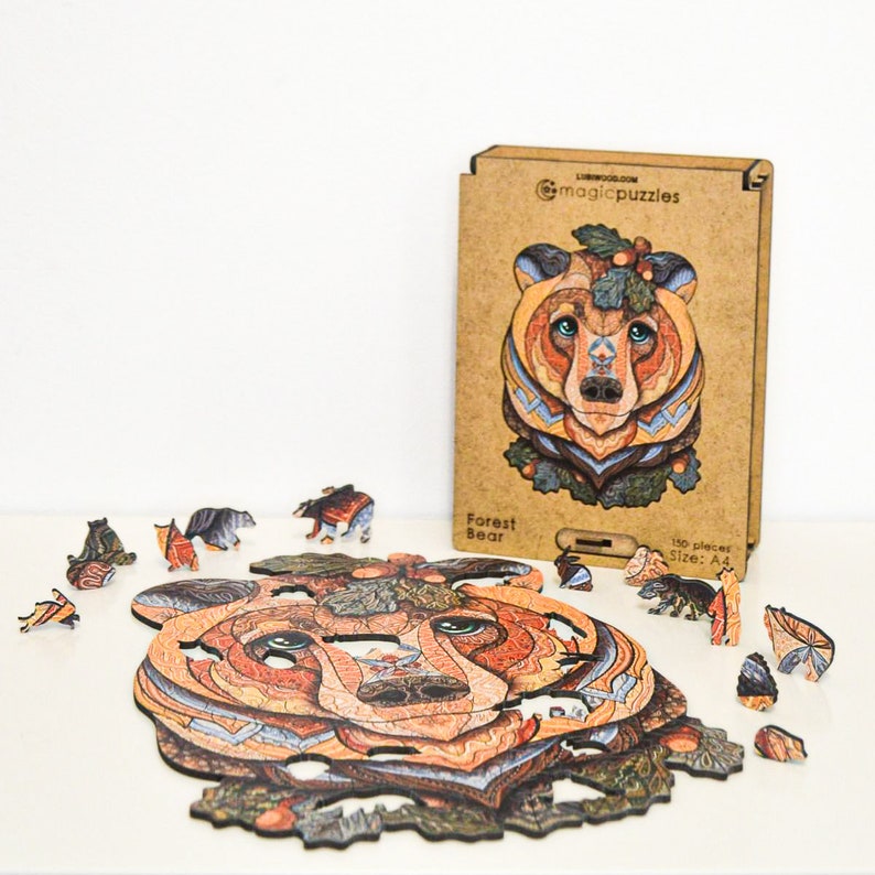May include: A wooden jigsaw puzzle featuring a detailed bear design, with pieces scattered around the partially assembled puzzle. The box shows the completed image and the text "Forest Bear" and "magicpuzzles". The puzzle has 150 pieces.