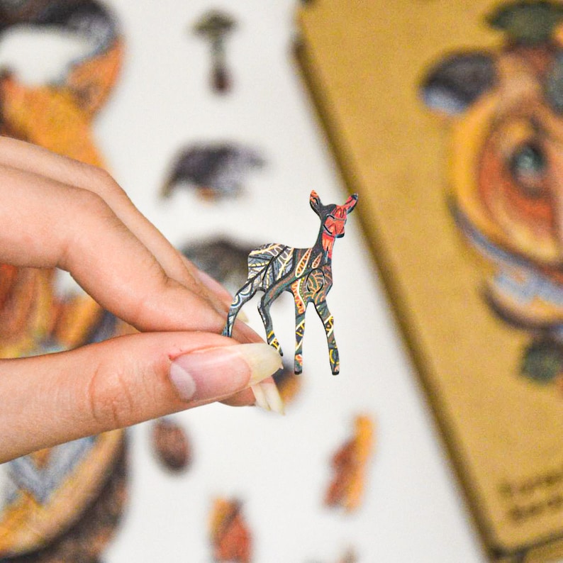 May include: A small, wooden deer figurine with a colorful, patterned design. The deer has a red face and is held between fingers. The figurine is part of a set of animal-themed wooden puzzles or decorations.