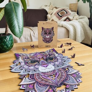 May include: A purple and grey wooden jigsaw puzzle of a cat's face with intricate details. The puzzle is assembled on a wooden table with a few pieces missing. The box for the puzzle is in the background and says "Curious Cat" on it.