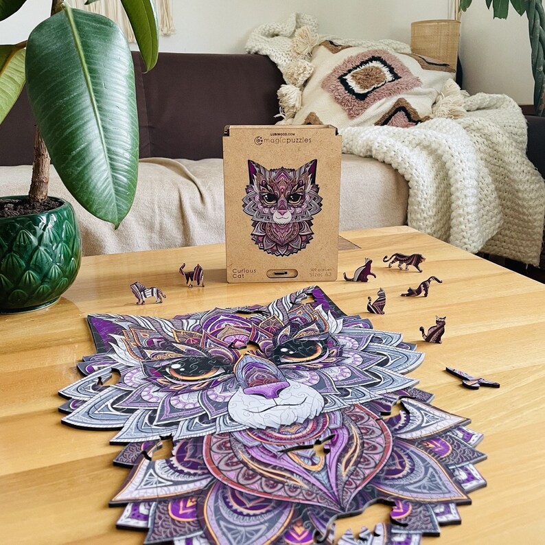 May include: A purple and grey wooden jigsaw puzzle of a cat's face with intricate details. The puzzle is assembled on a wooden table with a few pieces missing. The box for the puzzle is in the background and says "Curious Cat" on it.