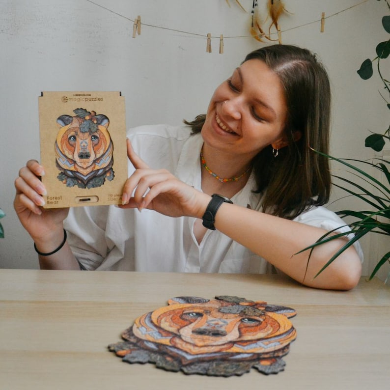May include: A wooden jigsaw puzzle featuring a bear design, with the text "Forest Bear" on the box. The puzzle is mostly shades of brown, orange, and blue. The box is held by a person.