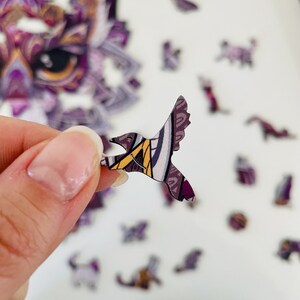 May include: A purple and yellow hummingbird puzzle piece with intricate patterns. The piece is made of cardboard and has a glossy finish.