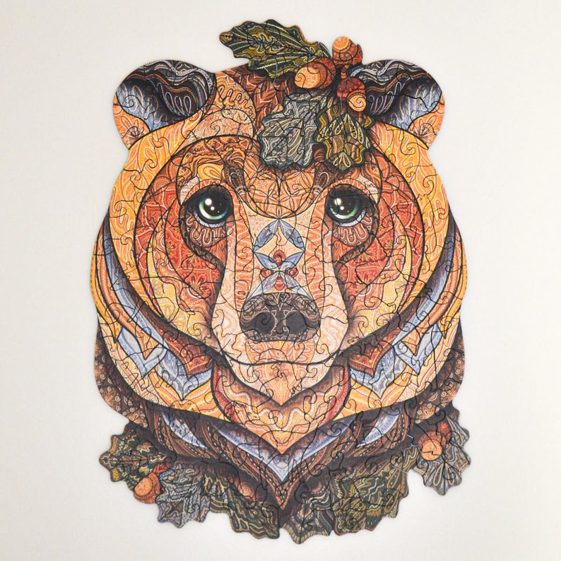 May include: A wooden jigsaw puzzle in the shape of a bear's head. The puzzle pieces are intricately designed with warm colors like orange, brown, and yellow. The bear has green eyes and a decorative leafy crown. A unique and engaging puzzle.