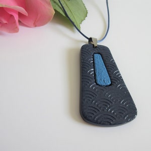 Puede incluir: Un collar con un colgante de cerámica azul oscuro y azul claro con un patrón de ondas. El colgante tiene forma de lágrima y cuelga de un cordón azul oscuro.