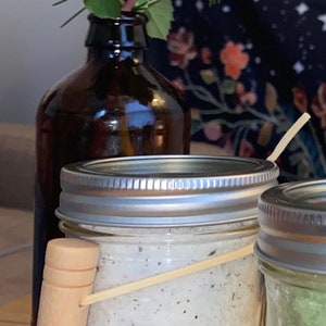 May include: Two glass jars with silver lids filled with bath salts. The jar on the left has a wooden scoop next to it. The jar on the right has green bath salts. The jars are sitting on a wooden surface.