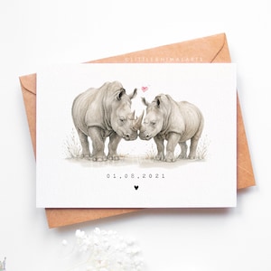 May include: A watercolour greeting card showing two rhinoceroses touching noses, with a small pink heart above them. The date "01.08.2021" is printed below the rhinos, with a black heart. The card is on a brown envelope.