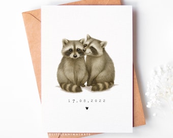 May include: A greeting card featuring two illustrated raccoons, one nuzzling the other. The date "17.08.2022" and a heart symbol are printed below the raccoons. The card is set against a brown envelope.