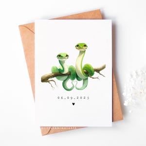 May include: A greeting card with two green snakes on a brown branch, tails intertwined in a heart shape. The card displays the date 06.09.2025 and a black heart symbol. The card rests on a brown envelope.
