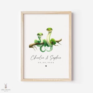May include: A framed print showing two green snakes on a branch. The names "Charlie & Sophie" are written in cursive below the snakes, along with the date 28.08.2026 and a heart. The artwork is in a watercolor style, with a light wooden frame.