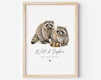 May include: A framed watercolor print featuring two raccoons. The raccoons are depicted in shades of gray and brown, with black markings around their eyes and striped tails. The print includes the names "Will & Sophie" and the date "20.07.2026".