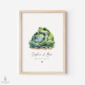 May include: A framed art print featuring a watercolor illustration of two green frogs embracing. The print includes the names "Sophie & Ben" and the date "07.06.2013" below the frogs, with a small heart symbol. The artwork is set against a cream background.