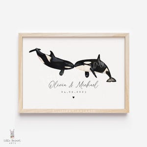 May include: A framed print showing two orcas, one black and white, touching noses. The print includes the names "Olivia & Michael" and the date "04.06.2025" in elegant script, with a small heart below. The frame is a light wood colour.
