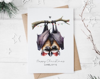 May include: A white greeting card featuring an illustration of two bats hanging upside down from a branch, wearing red and white Santa hats. The branch is decorated with colorful string lights. The text reads Happy Christmas CHARLOTTE.