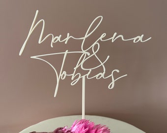 May include: White cake topper with the names "Marlena & Tobias" written in cursive script. The topper is on a white cake with pink flowers.