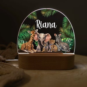 May include: A wooden night light with a clear acrylic insert featuring a jungle scene with a giraffe, elephant, zebra, lion cub, and a meerkat. The name "Riana" is written in white on the acrylic insert.