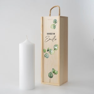 Könnte beinhalten: Eine Holzkiste mit einer weißen Kerze und einem grünen Eukalyptus-Design. Die Kiste hat einen Seilgriff und der Text "Taufkerze von Emilia" ist auf der Vorderseite aufgedruckt.