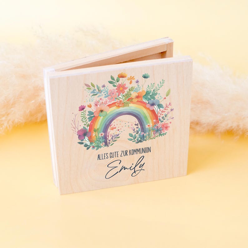 Peut inclure: Un coffret en bois clair avec un couvercle &agrave; charni&egrave;re, orn&eacute; d'un arc-en-ciel color&eacute; et de fleurs, avec le texte "ALLES GUTE ZUR KOMMUNION" et le nom "Emily".