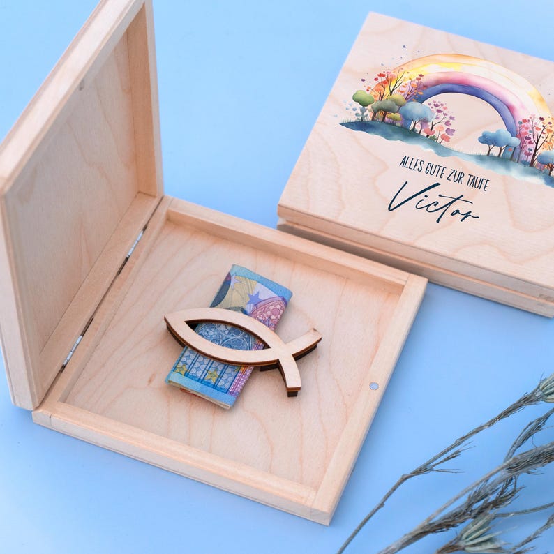 Peut inclure: Une bo&icirc;te en bois de couleur claire avec un couvercle ouvert, r&eacute;v&eacute;lant un symbole de poisson en bois et un billet pli&eacute;. Le couvercle de la bo&icirc;te pr&eacute;sente un motif d'arc-en-ciel &agrave; l'aquarelle avec le texte "Alles Gute zur Taufe Victor".